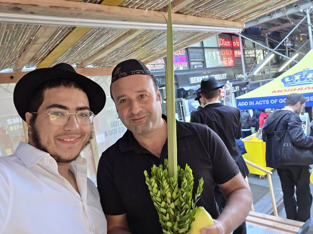 א יונגער חב״ד בחור מיט א היטל מאכט א סעלפי מיט א שמייכלענדיגן מאן וואס האלט א לולב און אתרוג אין א סוכה-מאביל אין מידטאון מאנהעטן.